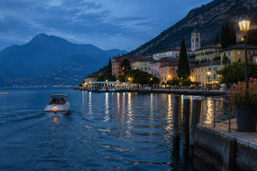 A small boat crossing calm evening water toward a lakeside town at dusk