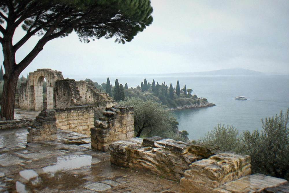 Stone ruins and a large pine tree overlook Lake Garda with a cypress-lined peninsula and a small boat under grey skies.