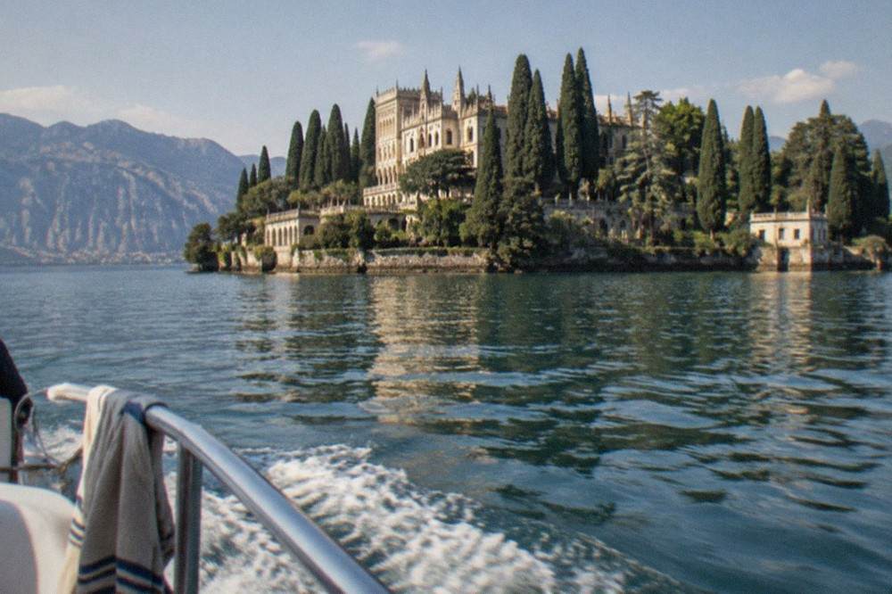 From a boat railing, a palatial stone building and tall cypresses sit on a small island in Lake Garda, with mountains behind and a wake in the water.