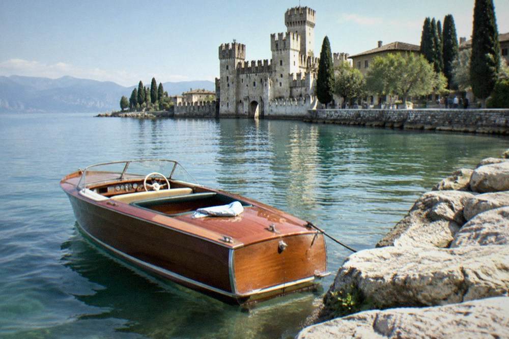 A classic wooden boat on clear green water near a Lake Garda town with a stone tower on the shore
