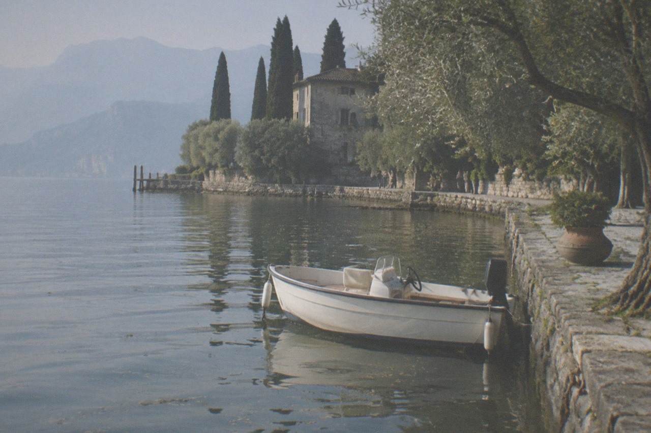 A small open motorboat moored in a Lake Garda harbour with olive trees and a stone waterfront in the background