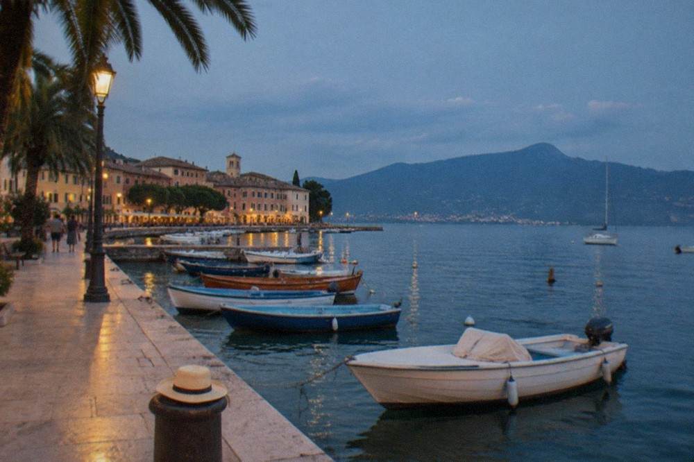 A lakeside promenade with palm trees, moored small boats and pastel buildings along the shore