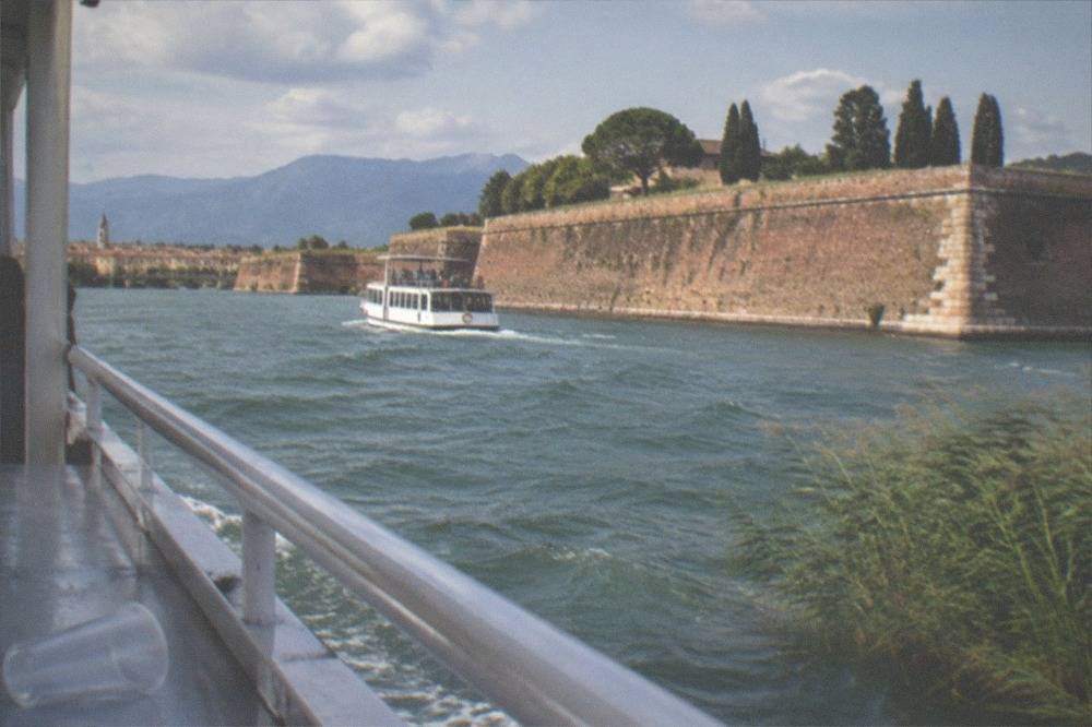 From a boat railing, a white passenger boat moves along water beside a high stone wall with trees, mountains and a town beyond, on Lake Garda.