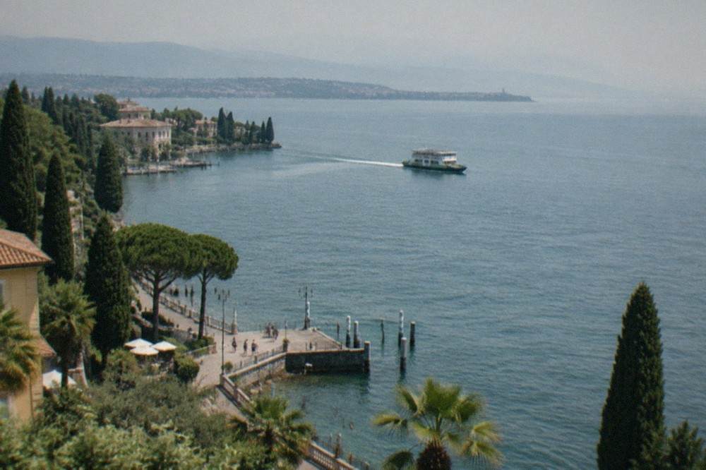 A long mountain lake seen from a high shore road, with a small town and ferry wake on the water below