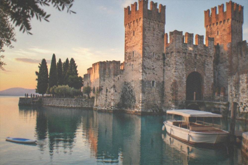 A tour boat moored beside a medieval castle at the tip of a lake peninsula under a clear sky