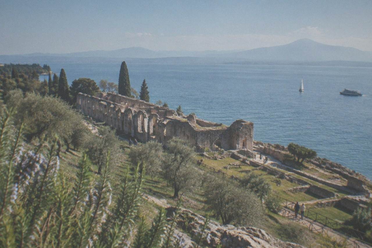 Stone ruins on a grassy hillside above a wide lake, with cypress and olive trees, distant mountains, a sailboat and a motorboat, on Lake Garda.