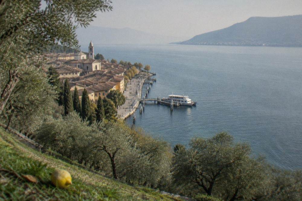 Hillside view of a Lake Garda lakeside town with a church tower, waterfront promenade and a ferry at a pier, olive trees in the foreground and mountains beyond.