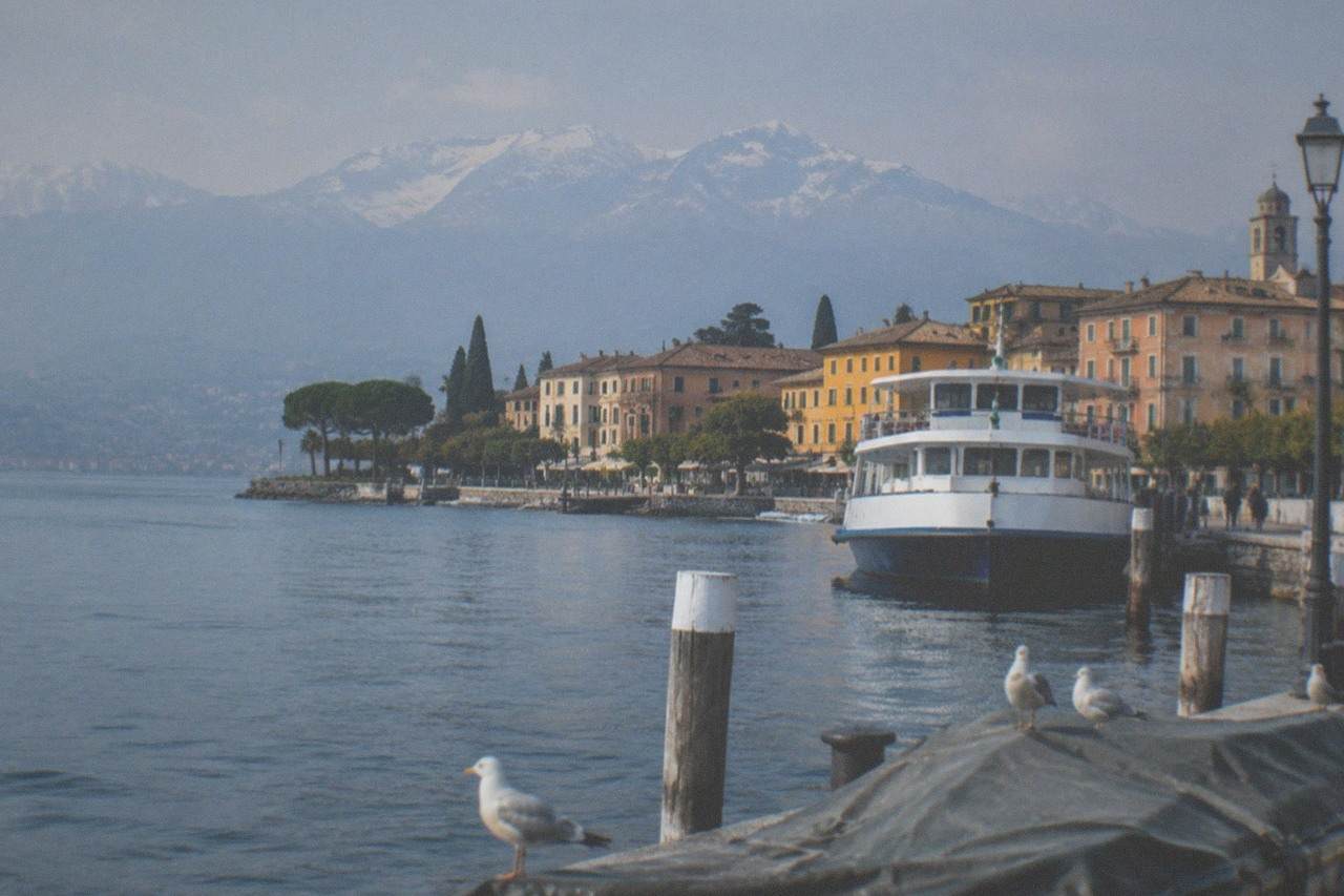 Lakeside quay with a white passenger boat, pastel buildings, snow-capped mountains beyond, and seagulls on wooden posts in the foreground, on Lake Garda.