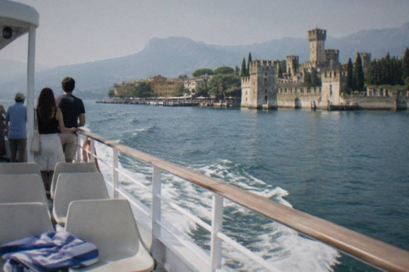 People stand at a boat railing looking toward a Lake Garda lakeside castle with towers, cypress trees and mountains beyond, wake trailing in the water.