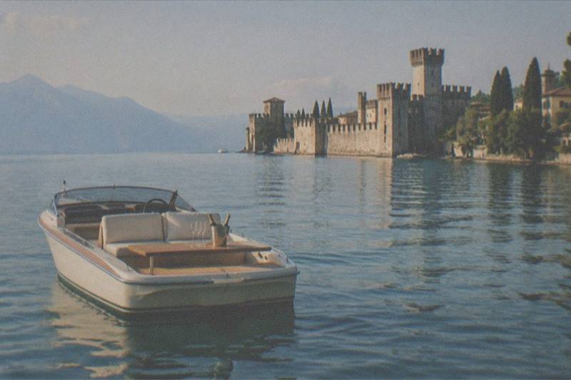 An empty white motorboat sits on calm water in the foreground, with a stone castle and towers on the shore and mountains beyond, on Lake Garda.