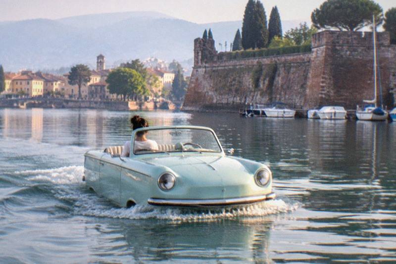 A pale blue vintage amphibious car with one person aboard cuts a wake across calm water beside a stone fortress, town and mountains, on Lake Garda.
