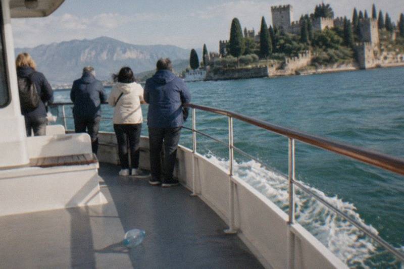 Four people stand at a boat railing looking toward a Lake Garda lakeside castle with towers and cypress trees, mountains beyond, a bottle on the deck.