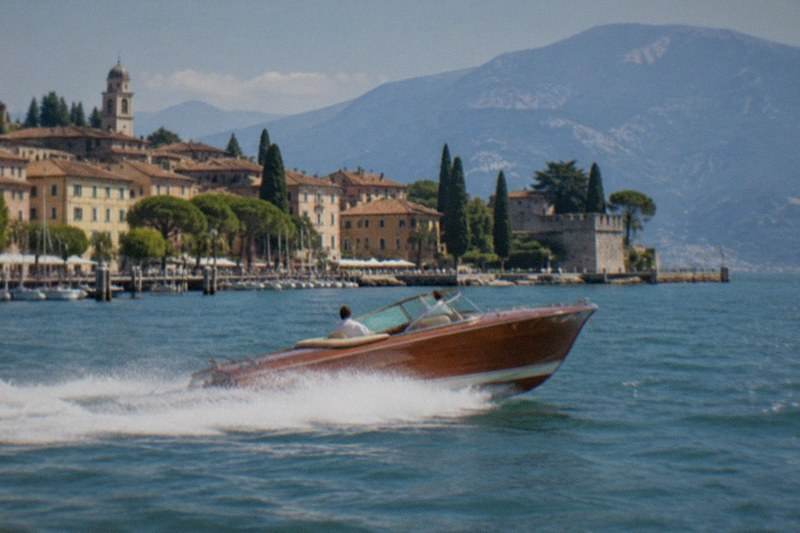 A gleaming vintage speedboat with polished wood trim carves a curve across Lake Garda, spray lifting at the bow, a skipper at the helm and steep hillsides rising behind in Italy.