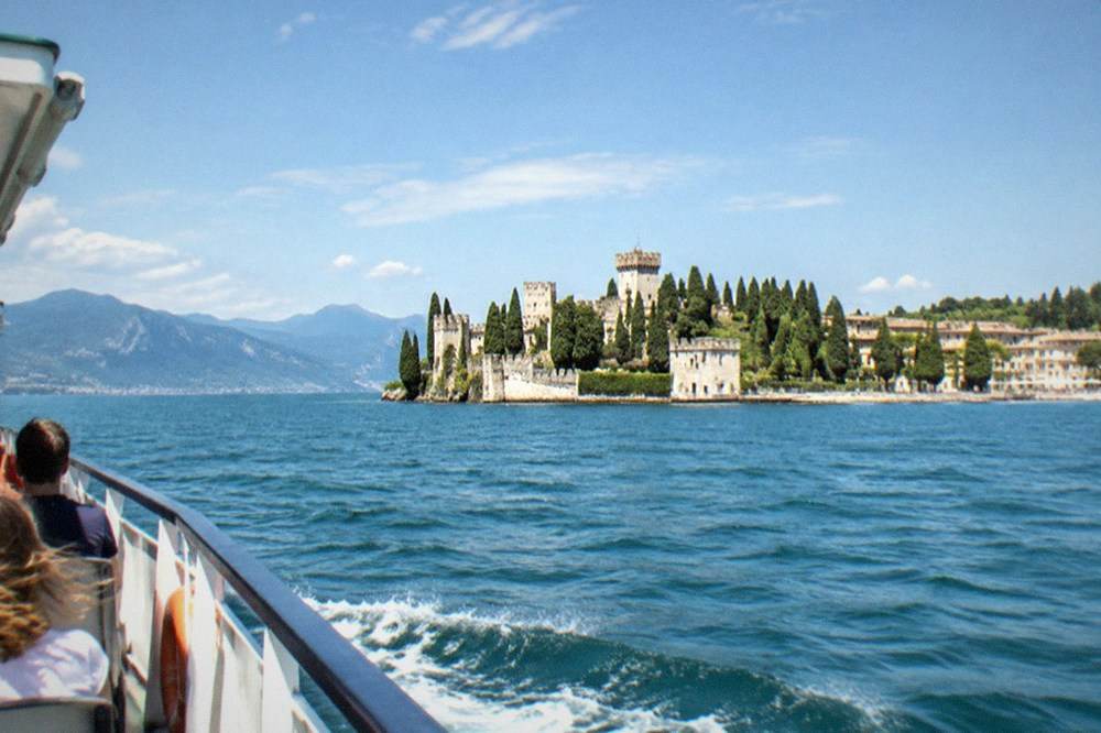 From a boat, a stone castle with towers and cypress trees sits on a Lake Garda lakeside peninsula, mountains beyond under a blue sky.
