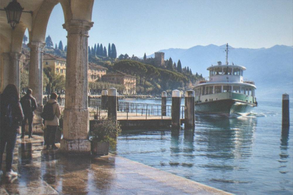 Stone arcade on a wet Lake Garda lakeside walkway with people, a wooden pier, and a ferry approaching, hills and buildings beyond.