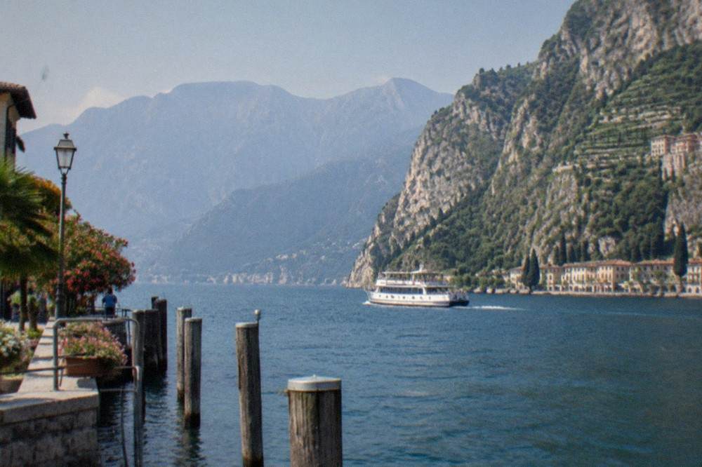 A small passenger ferry crossing open water toward the cliffs of the northern Lake Garda shore