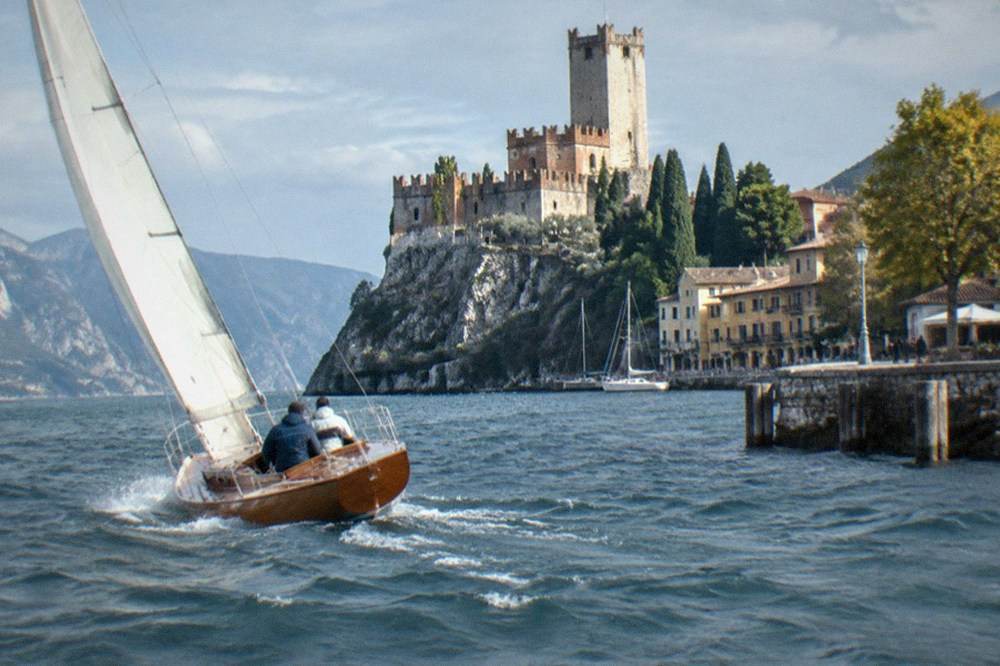 A historic sailboat heeling gently on open water near Malcesine with a stone castle on the headland