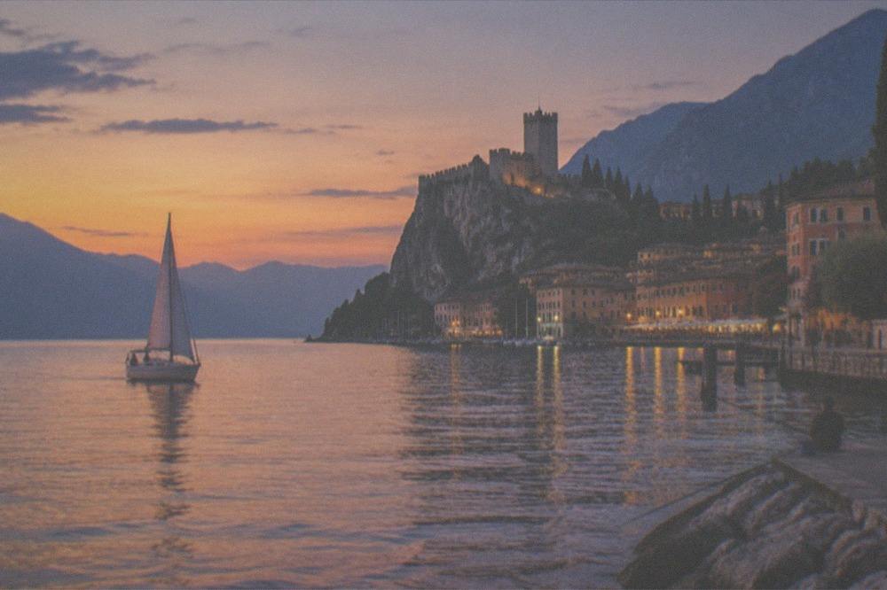 An evening sailboat on calm golden water near the Malcesine headland with the town lit behind