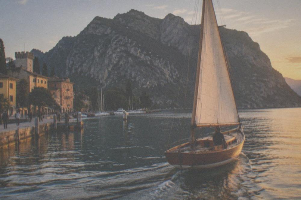 A wooden sailboat with a tall white sail glides near a Lake Garda lakeside promenade and buildings, with a steep mountain behind.