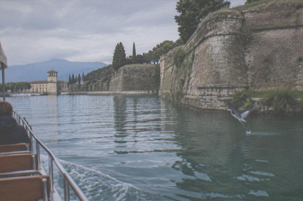 A low boat passing close to a rocky peninsula tip with cypress trees and a distant castle on the shoreline