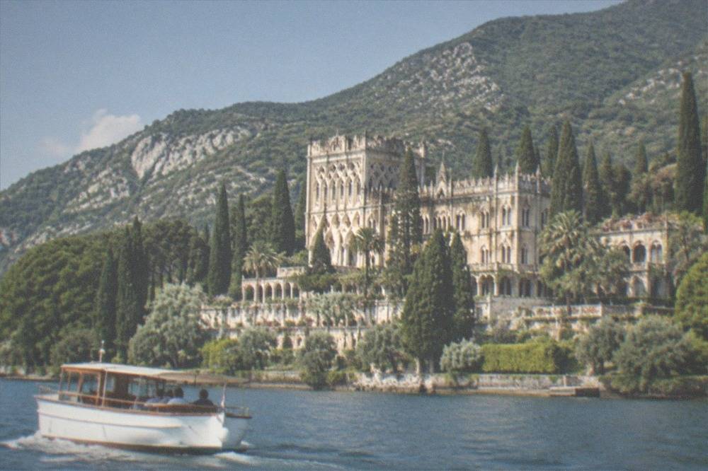 A small tour boat cruising past a castle on a rocky peninsula jutting into Lake Garda