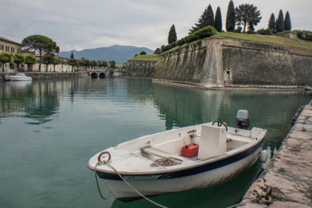 A small white open motorboat with a blue stripe is tied to a stone quay on calm water, with fortress walls, trees and mountains under cloudy skies, on Lake Garda.