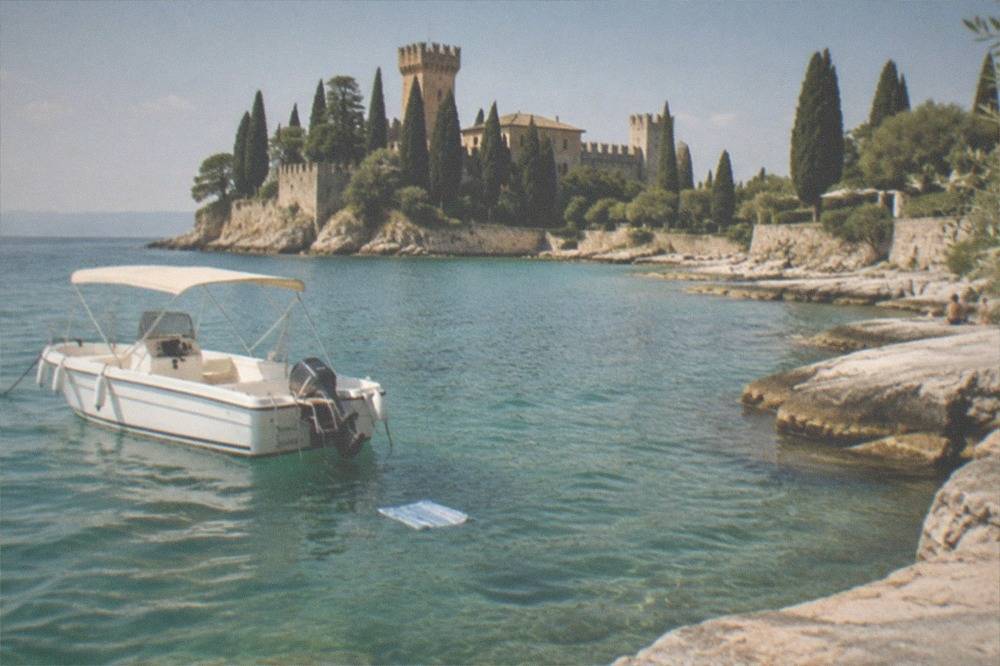 A white motorboat with a beige canopy floats in clear turquoise water beside a rocky shore, with a castle and cypress trees on a peninsula behind, on Lake Garda.
