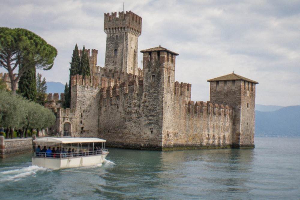 A small shared tour boat passing the fortified walls and towers of a lakeside castle in Sirmione