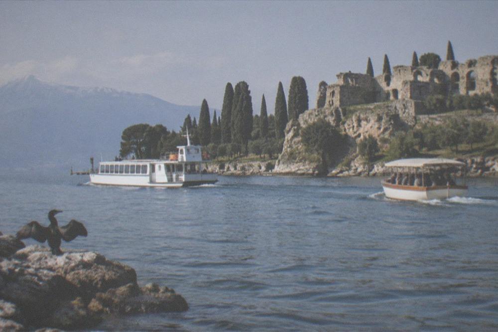 A ferry and a small tour boat crossing the same stretch of Lake Garda water near a rocky headland