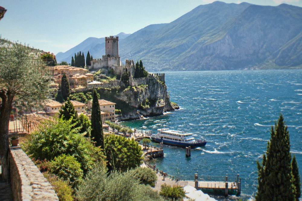 Lakeside village and stone castle on a cliff, with a ferry at a wooden pier, cypresses, and mountains across choppy blue water, on Lake Garda.
