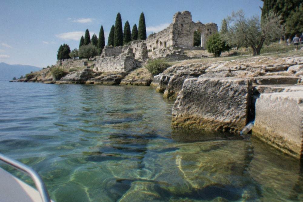 Clear Lake Garda water against large stone blocks on a rocky shore, with ruined walls and tall cypress trees under a blue sky.