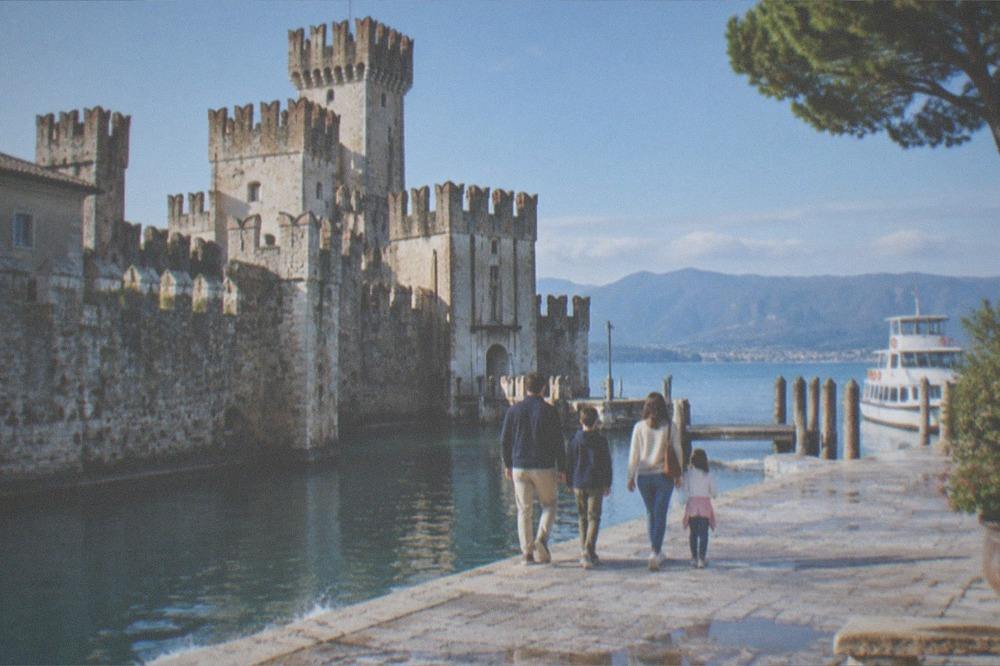 A family of four walks away along a stone Lake Garda lakeside promenade toward a medieval castle, with a docked boat to the right and mountains beyond.