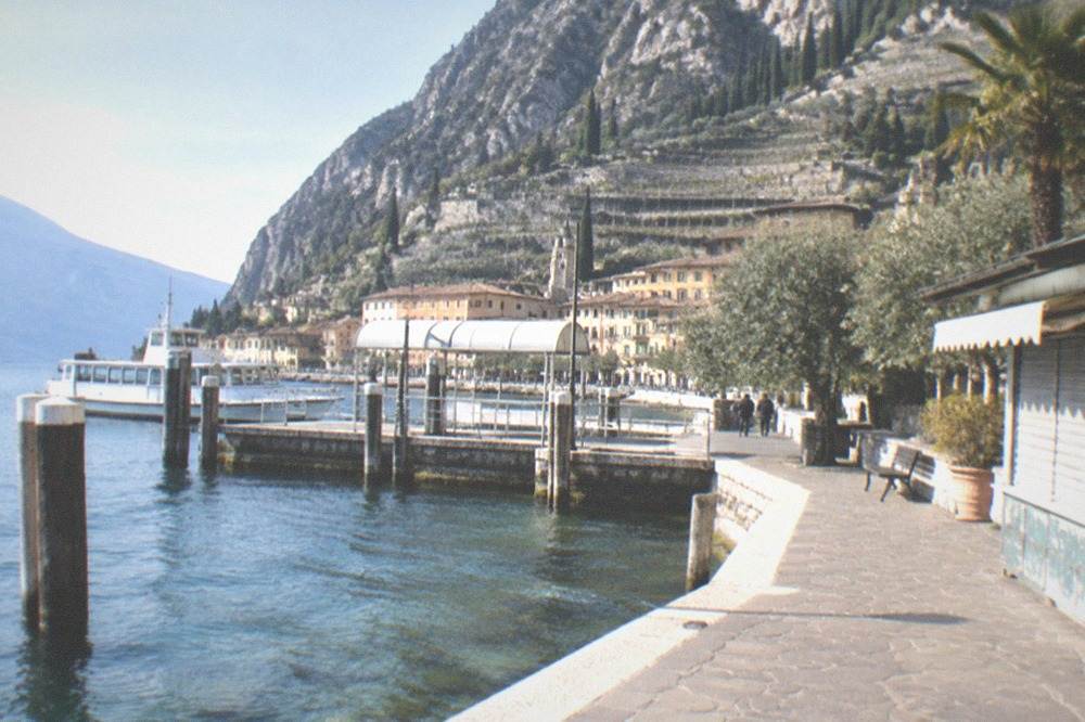 Lakeside stone promenade with a docked ferry at a pier, hillside town and mountains, a few walkers, and a shuttered kiosk, on Lake Garda.