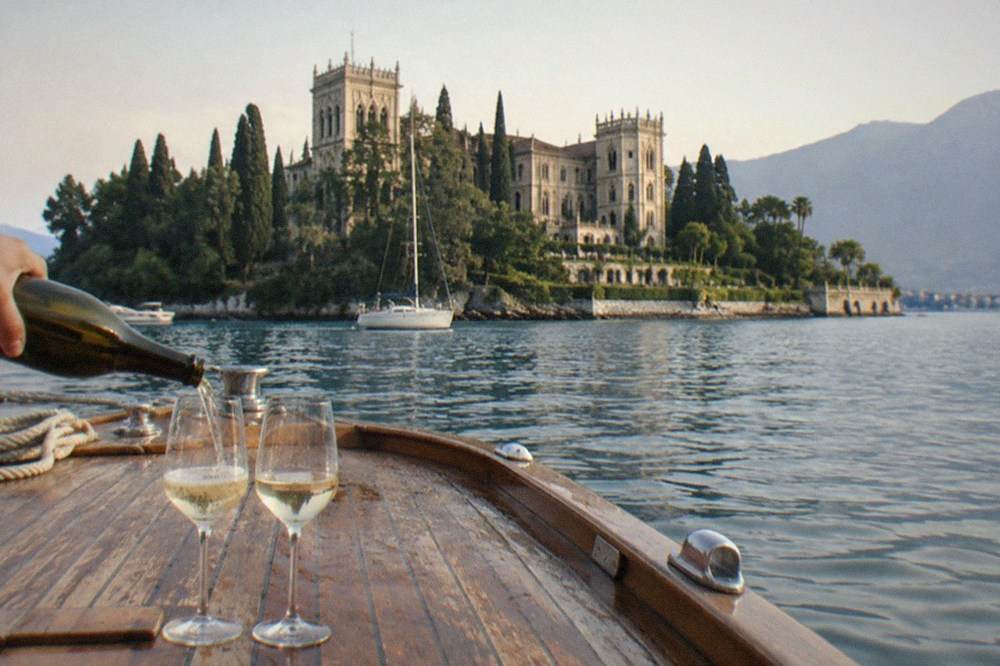 A hand pours white wine into two glasses on a wooden boat deck, with a turreted villa, cypresses and a sailboat on Lake Garda behind.