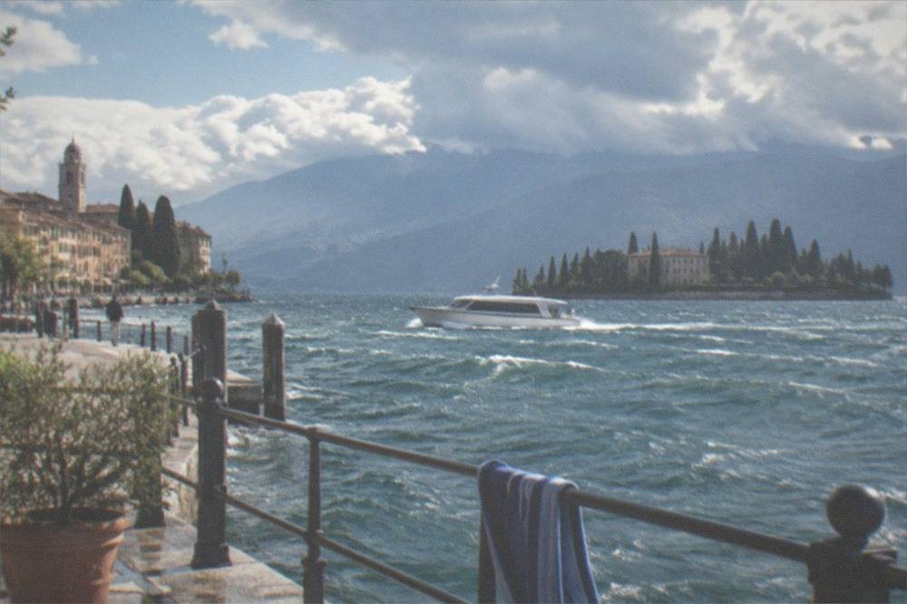 From a Lake Garda lakeside railing with a potted plant and towel, a white motorboat moves on choppy water past a town clock tower toward a tree-lined island and mountains.