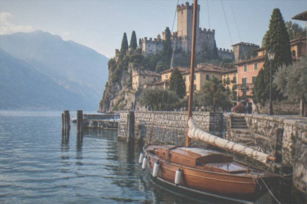 A wooden historic sailboat moored at a stone harbour wall in a Lake Garda town
