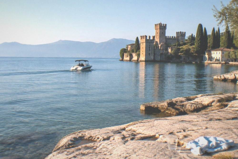 A motorboat on calm water near a rocky shore, with a Lake Garda lakeside castle, cypress trees, distant mountains, and a glass and cloth on the rocks.