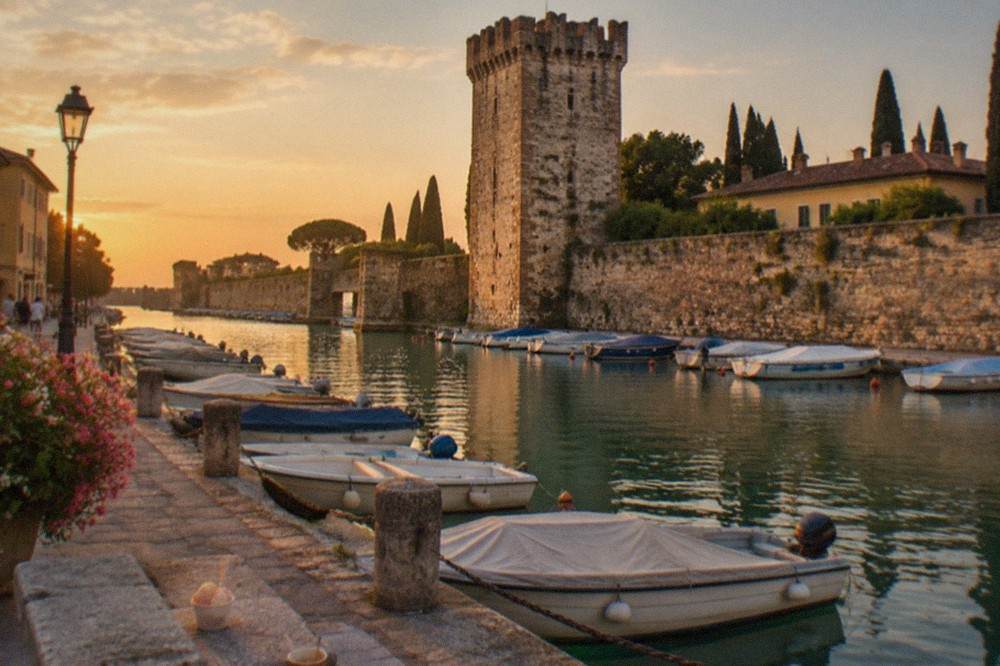 A southern Lake Garda harbour at golden hour with a stone castle tower above moored small boats and a lakeside promenade