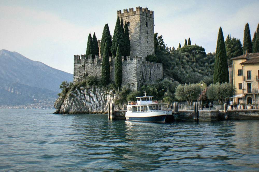 An eastern shore harbour on Lake Garda with a stone castle on a rocky outcrop, cypress trees and a small ferry at its mooring