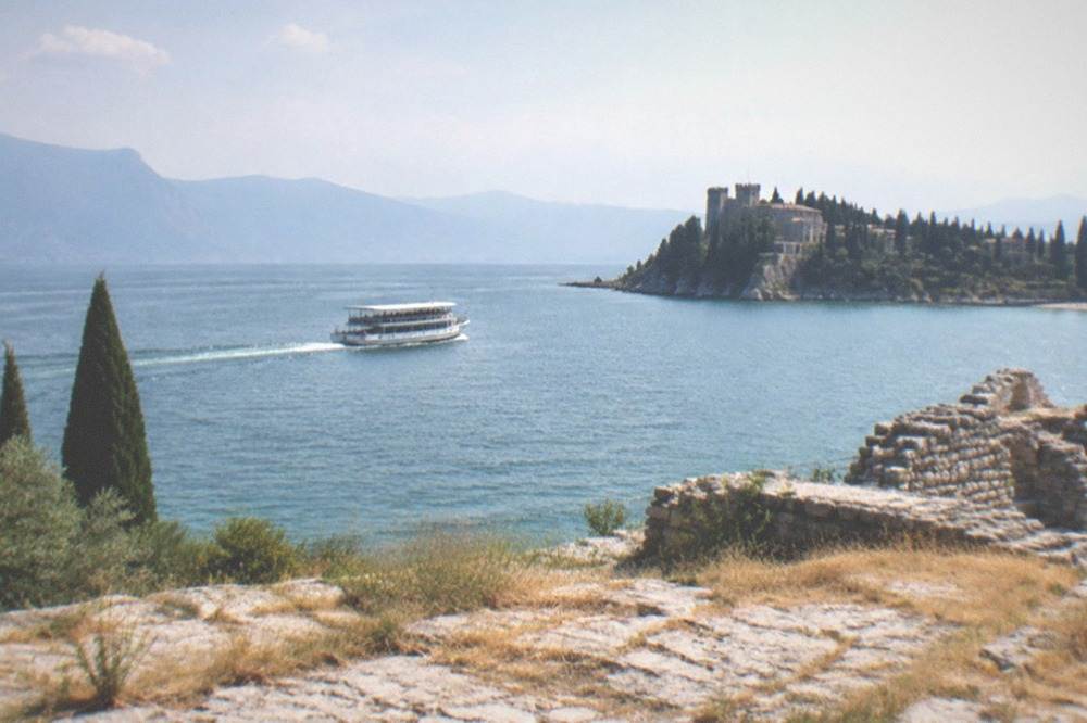 A white passenger boat on Lake Garda heads toward a wooded peninsula with a castle, cypress trees and stone ruins in the foreground, mountains beyond.