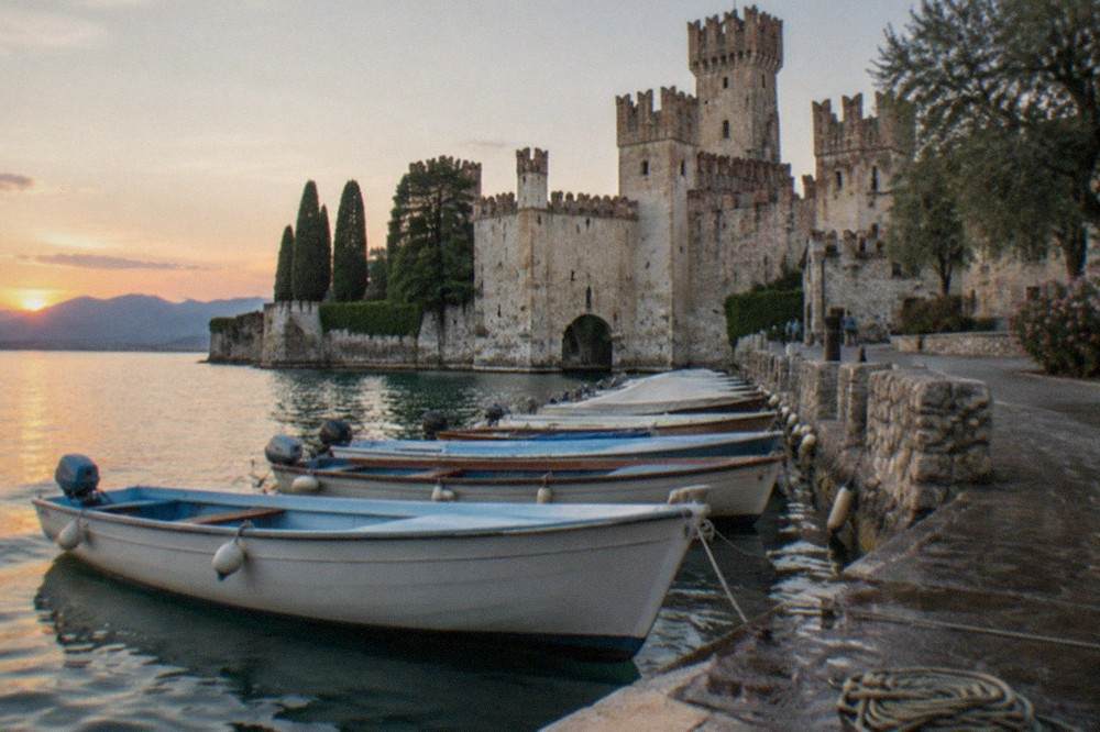 A row of small open passenger boats moored along a stone harbour wall beneath a medieval castle