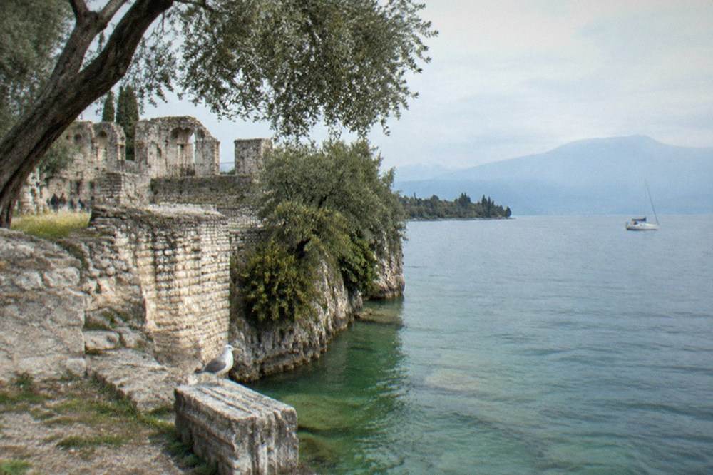Stone ruins and trees line a lakeshore with a seagull on a block, a sailboat on calm water and mountains beyond, on Lake Garda.
