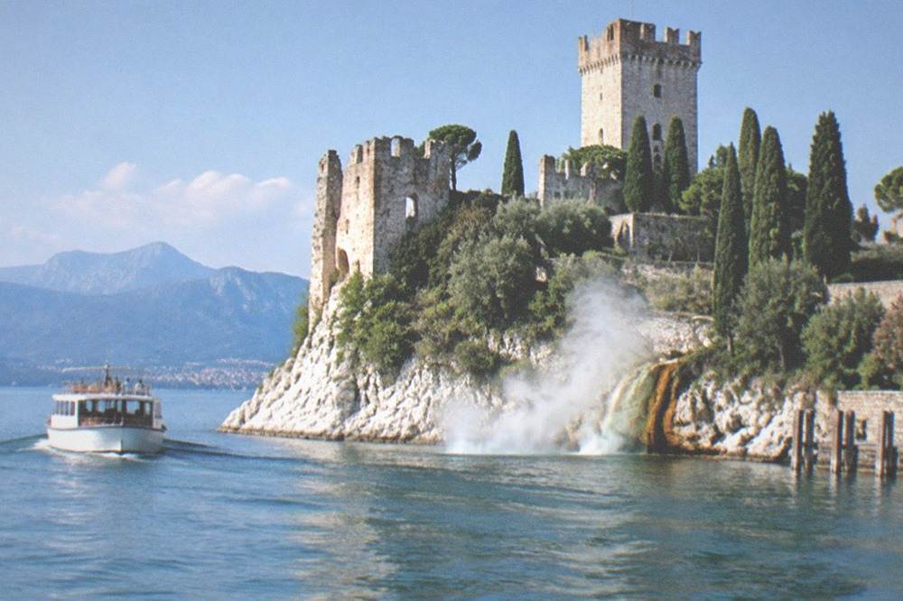 A square-towered castle on a rocky, tree-lined peninsula, a white passenger boat on Lake Garda, distant mountains, and spray at the cliff base.