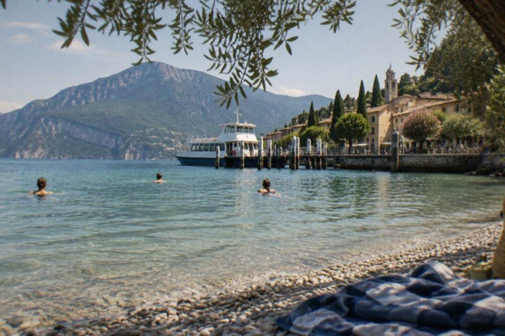 Swimmers in clear water beside a pebble beach with olive trees on the shore