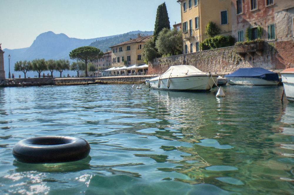 A black inner tube floats on rippled water beside moored boats, with Lake Garda lakeside buildings, trees and mountains in the background.