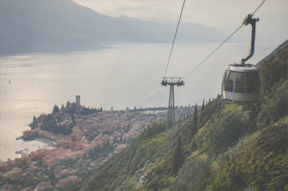 A cable car cabin ascending a steep green mountainside above a lake town
