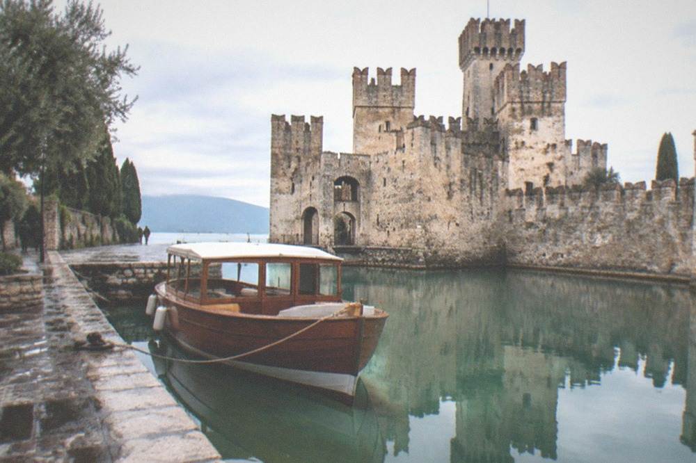 A wooden tour boat moored beside a stone castle wall reflected in still lake water