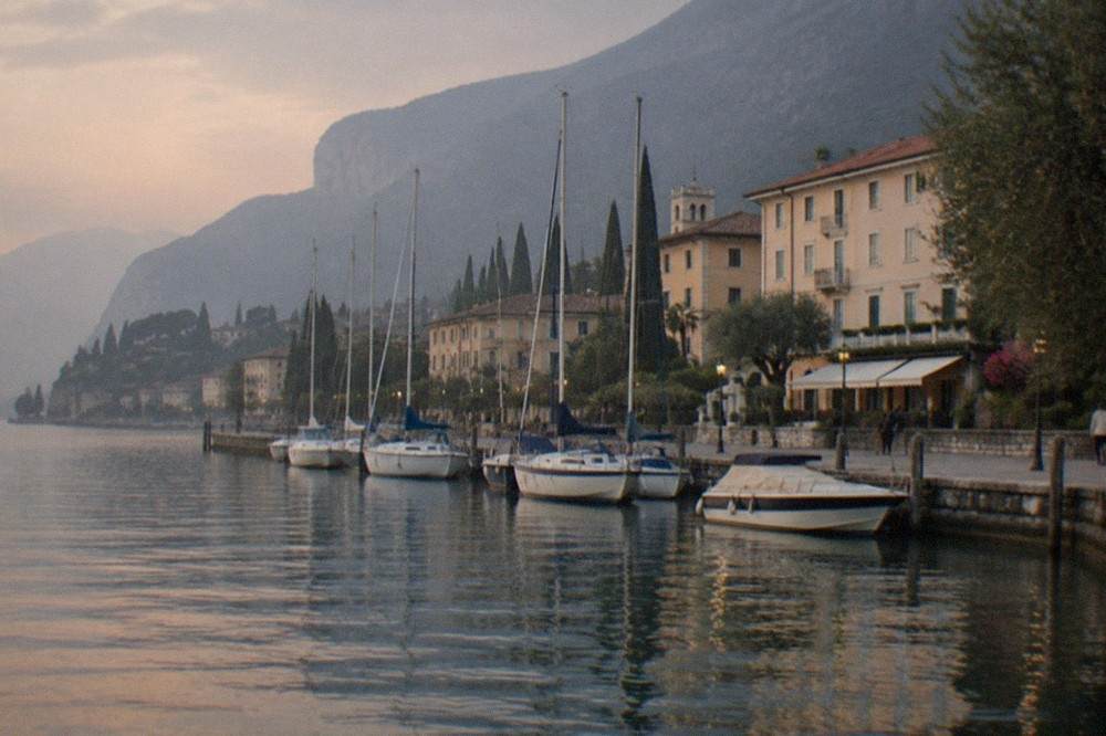 Sailboats and a motorboat moored along a stone quay beside Lake Garda lakeside buildings, with mountains beyond at dusk.
