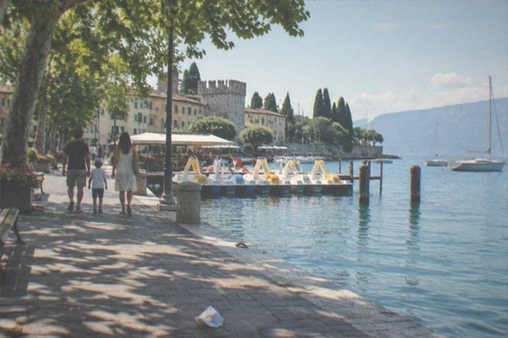 A family walks a tree-lined stone promenade beside Lake Garda with yellow-canopied pedal boats, sailboats and a towered waterfront.