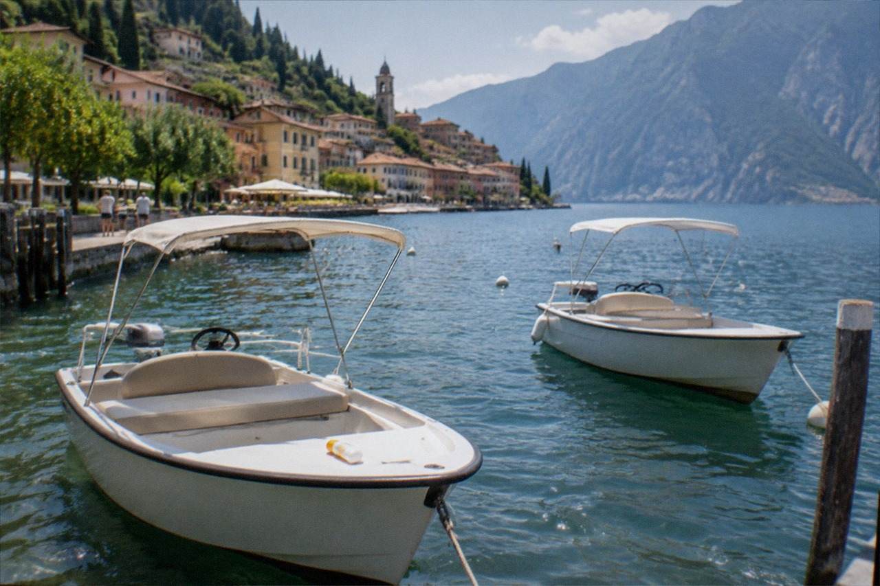 Two small white canopy boats moored at a Lake Garda lakeside promenade, with a hillside town, church tower and mountains across the water.